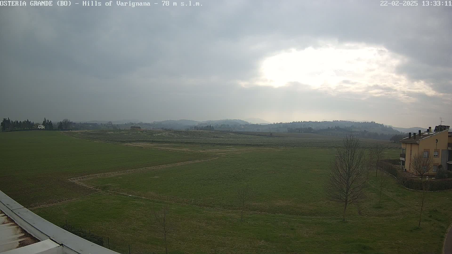 A mostly cloudy sky hangs over a flat expanse of green farmland, with a few buildings visible in the distance and a few leafless trees near the bottom right.