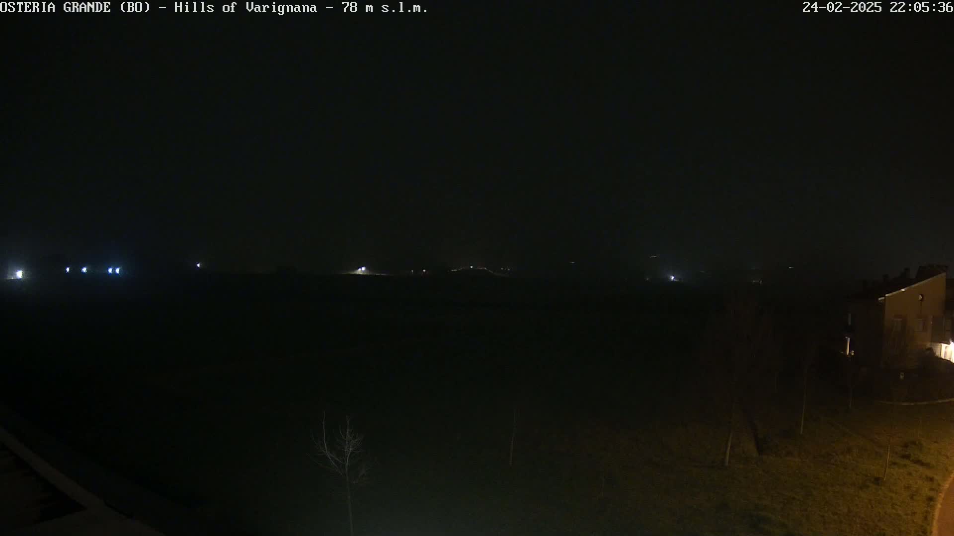 A dark, nighttime scene shows a mostly flat landscape with a few distant lights and a building visible in the lower right corner under a hazy sky.