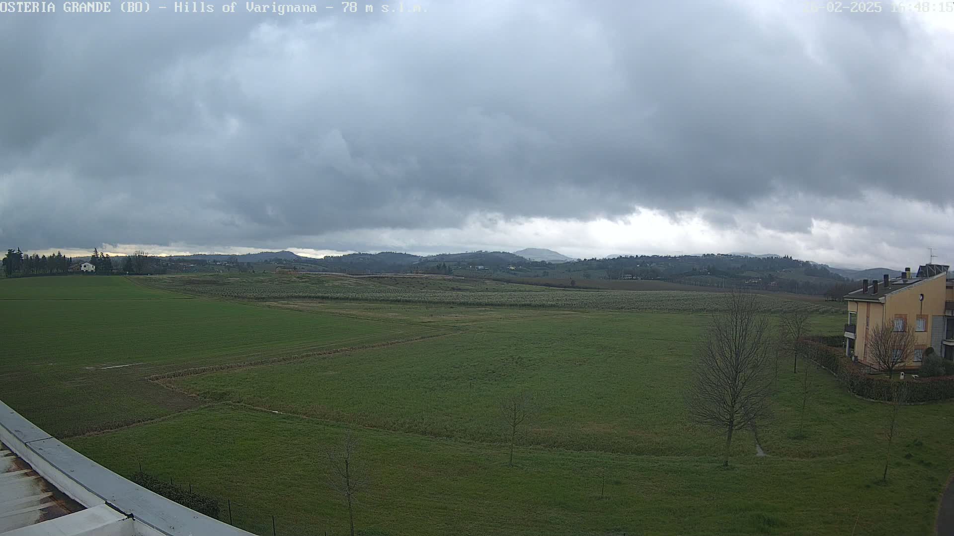 A wide, overcast landscape of green fields and distant hills, with a few buildings visible in the lower right corner.