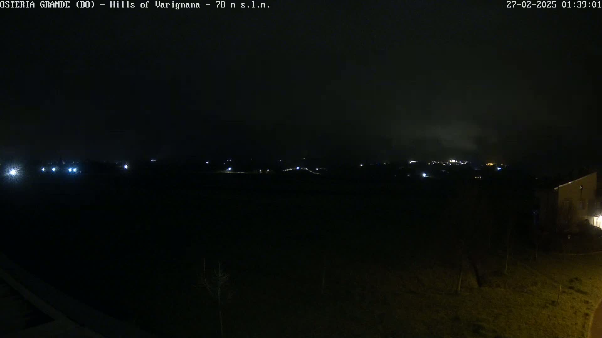 A nighttime, dark view of a landscape with distant lights and a building partially visible in the bottom right corner, under a mostly clear sky.