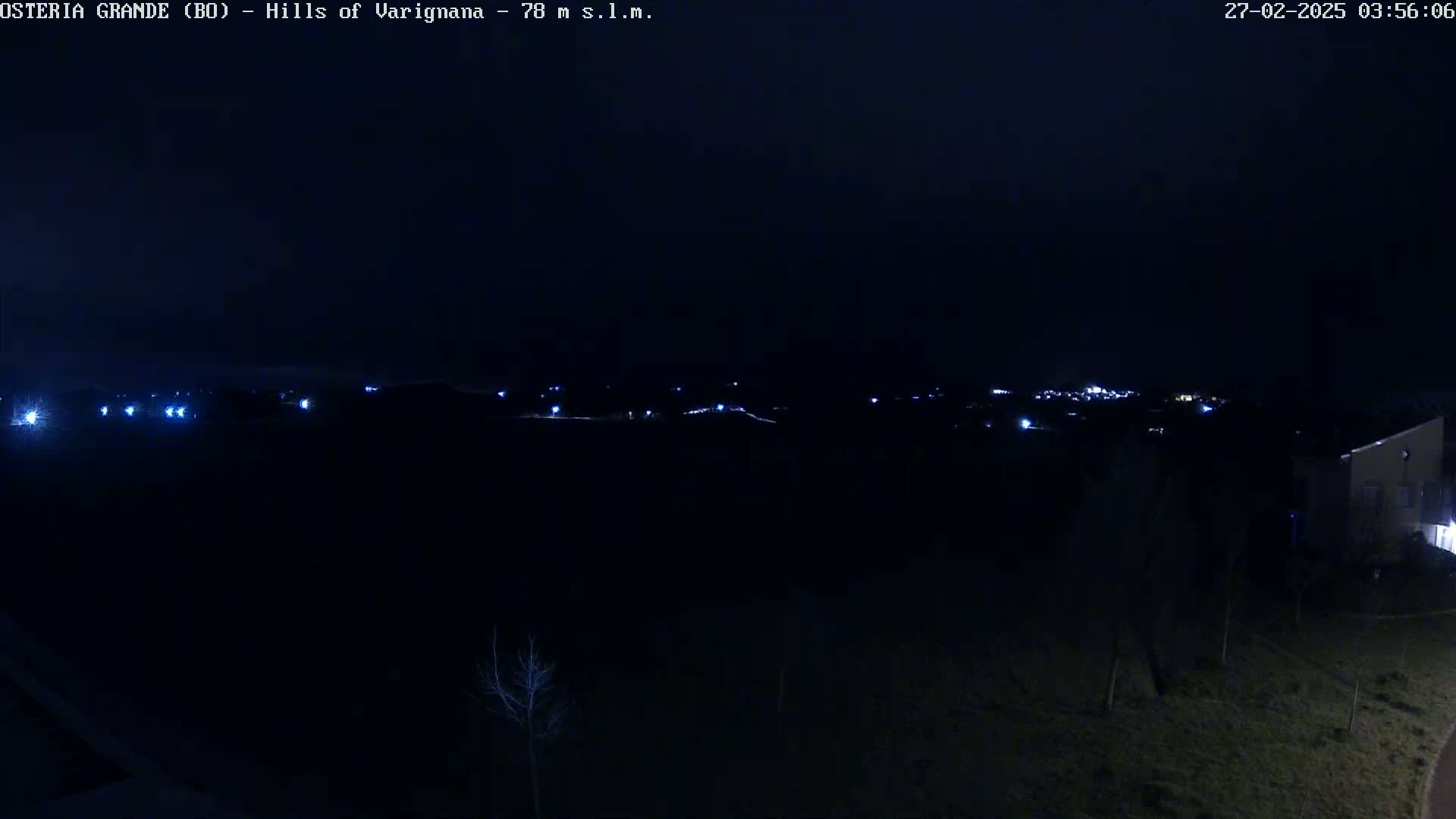 A nighttime, long shot of a dark landscape with a few distant lights and a building partially visible on the lower right.
