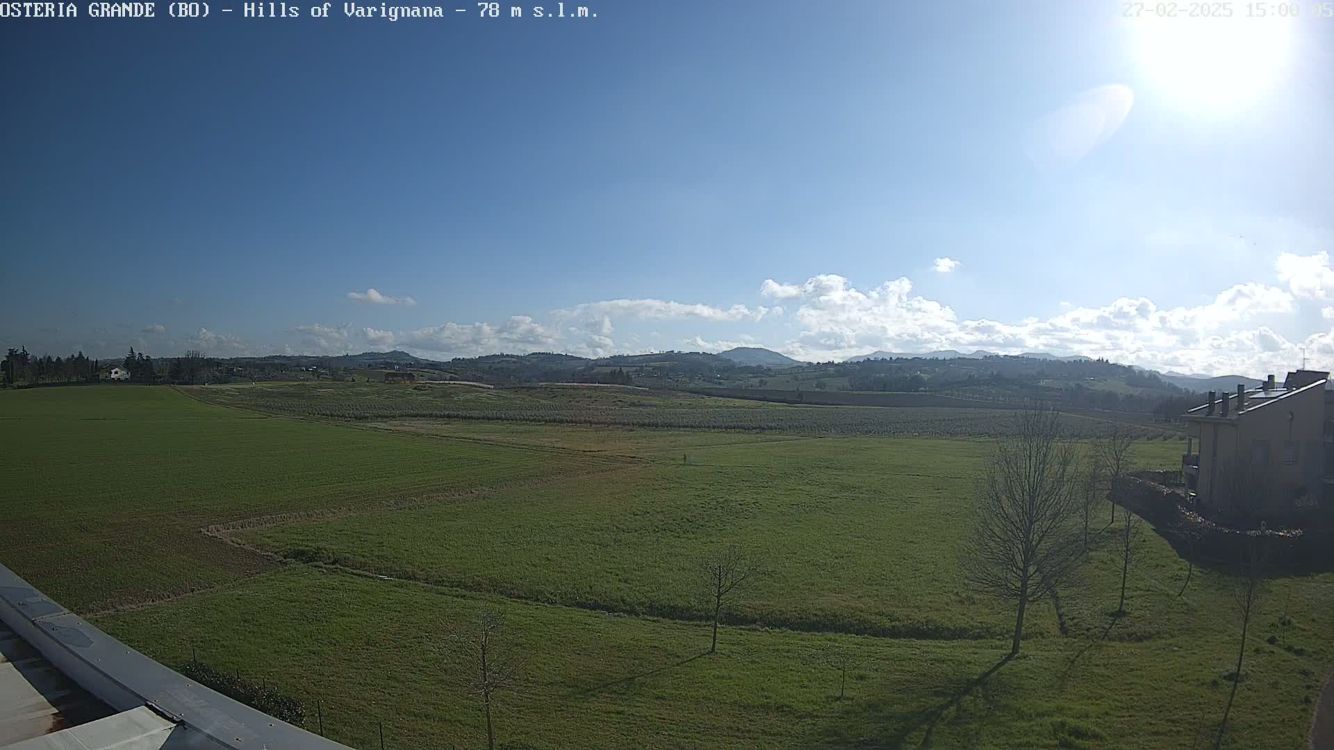 A sunny day with partly cloudy skies overlooks a vast expanse of green fields, a few scattered trees, and a building in the distance, all set against the backdrop of rolling hills.