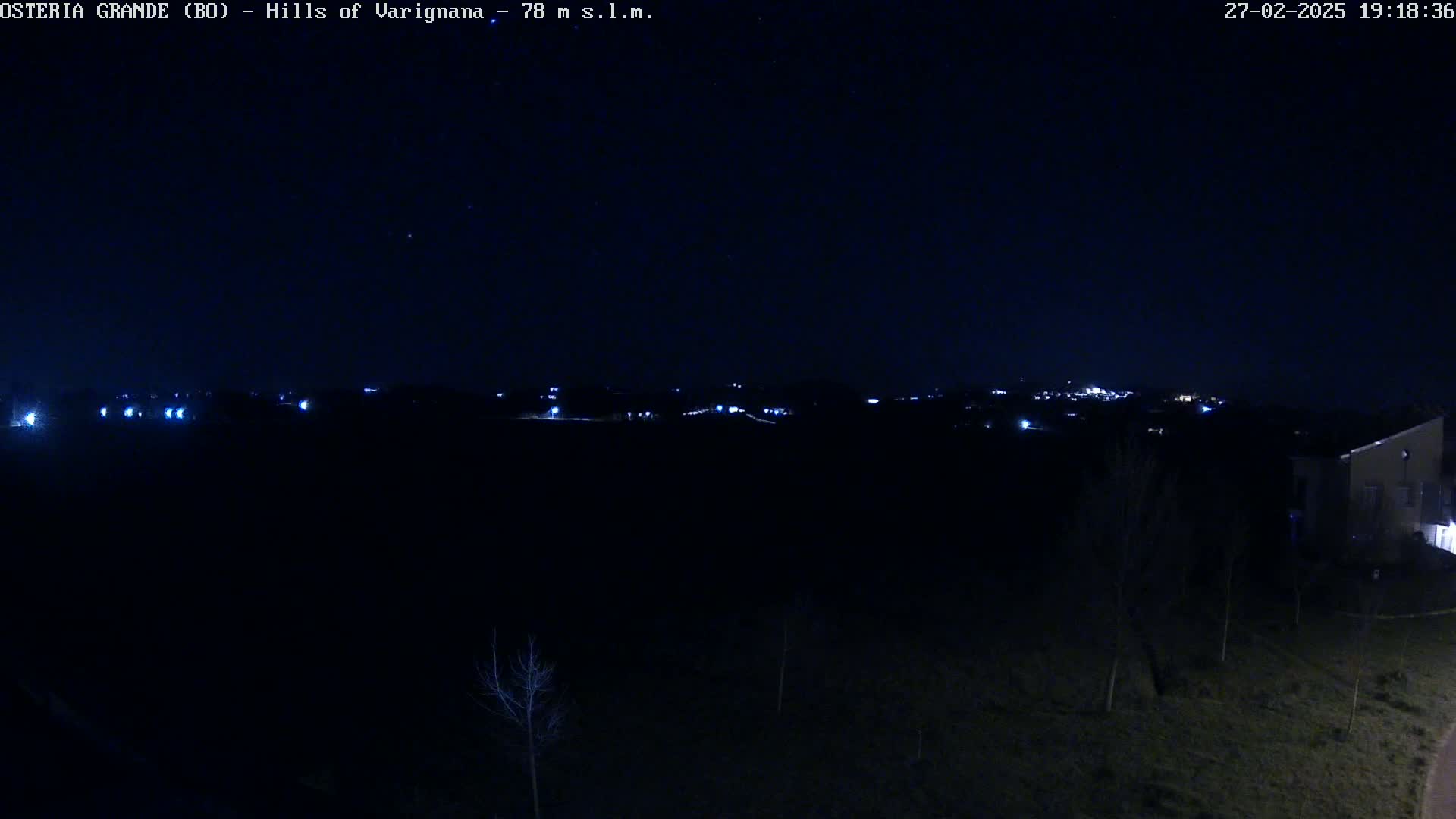 A nighttime, dark landscape shows a distant line of faint lights, a few bare trees, and a building in the lower right corner under a clear, starlit sky.
