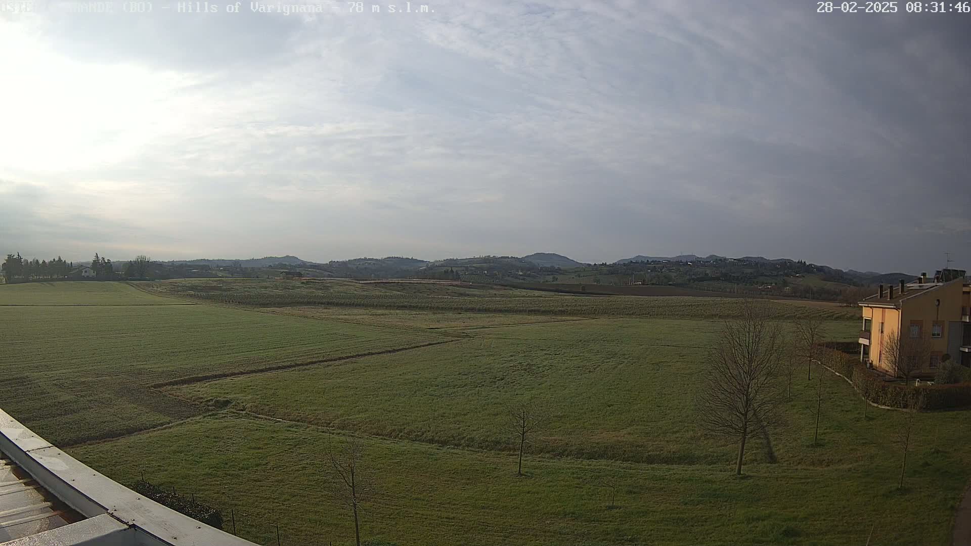 A wide expanse of green fields stretches towards a line of rolling hills under a mostly cloudy sky.