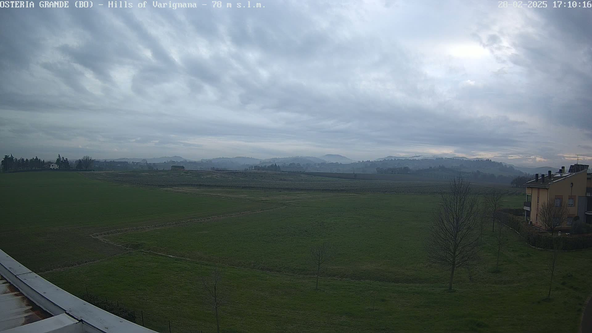 A flat green field stretches to the base of rolling hills under a cloudy sky.