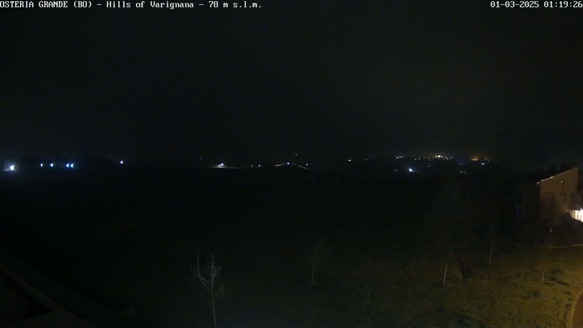 A nighttime, dark landscape view shows a few distant lights in the middle distance and a single building at the lower right.