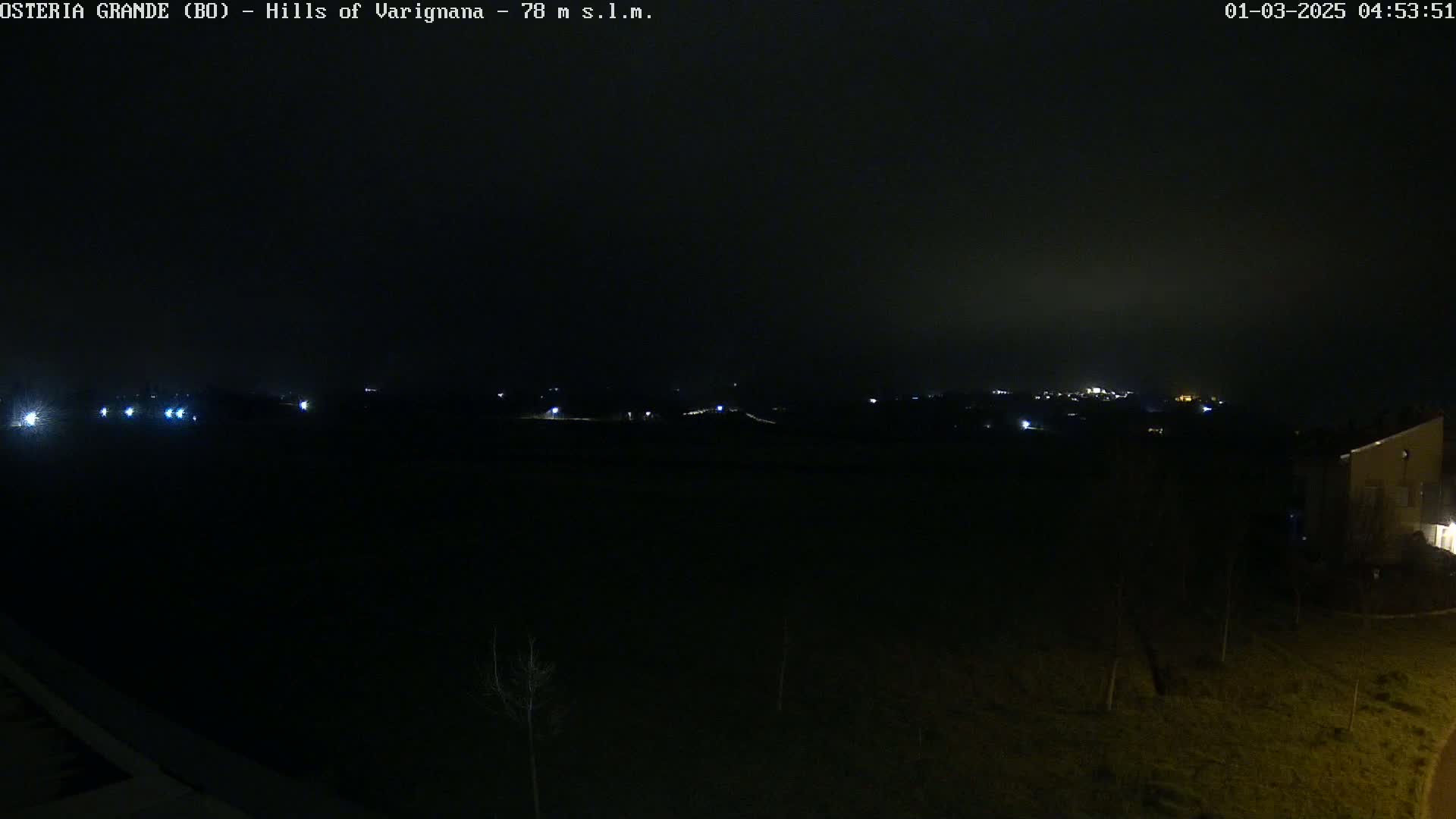 A dark, nighttime scene shows a distant, dimly lit town and a few lights scattered across a dark field, under a hazy sky.