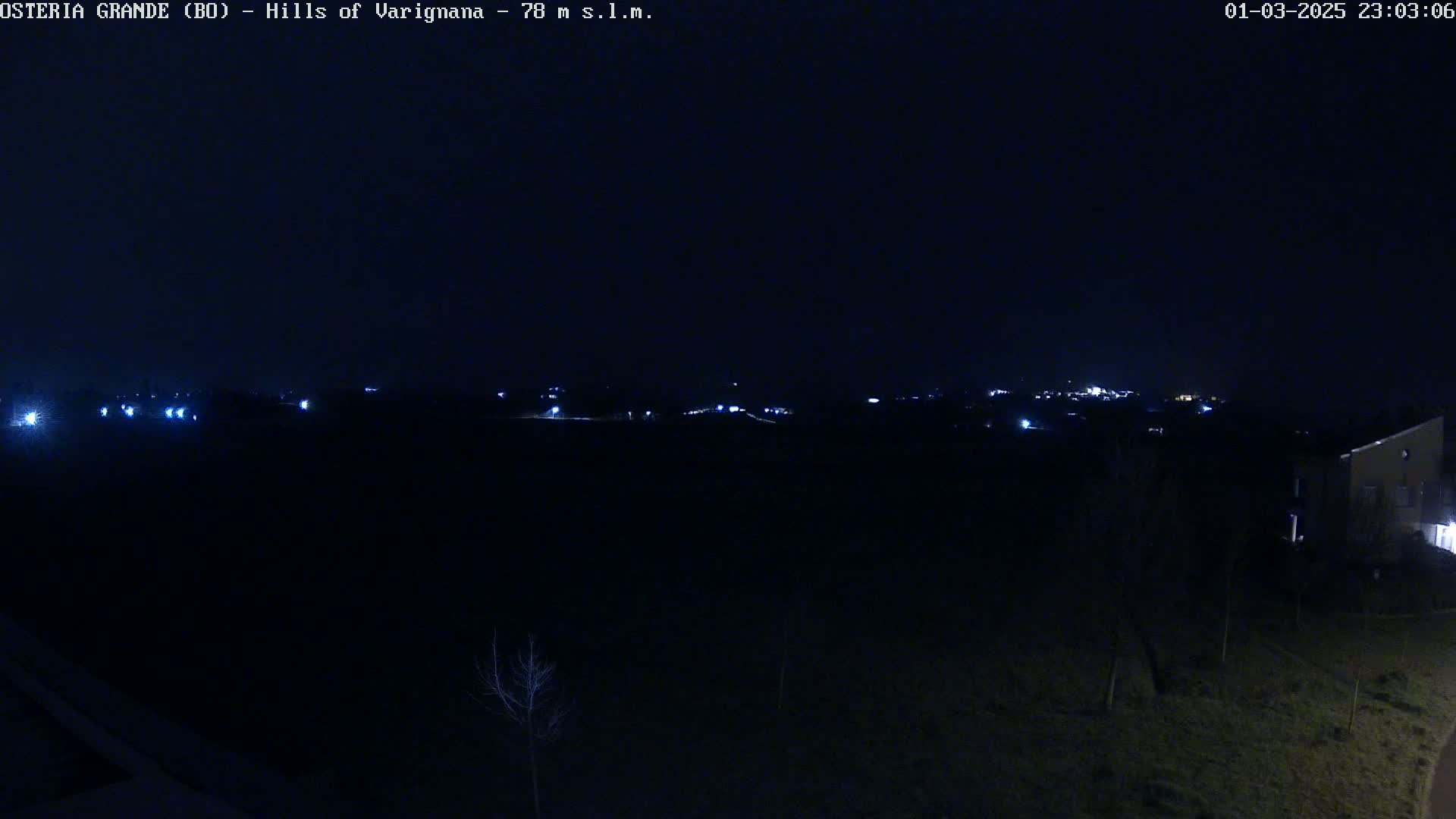 A dark, nighttime landscape shows a distant line of lights from a town or village, partially obscured by darkness.