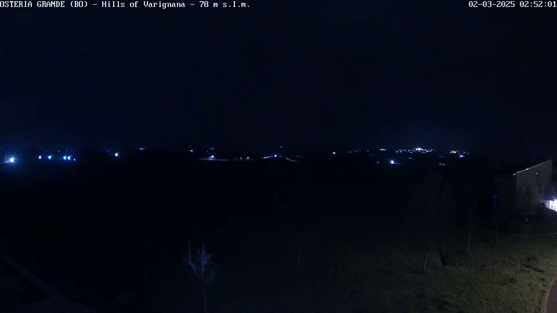 A nighttime view of a dark landscape with a few distant lights visible on a hill, under a clear, dark sky.