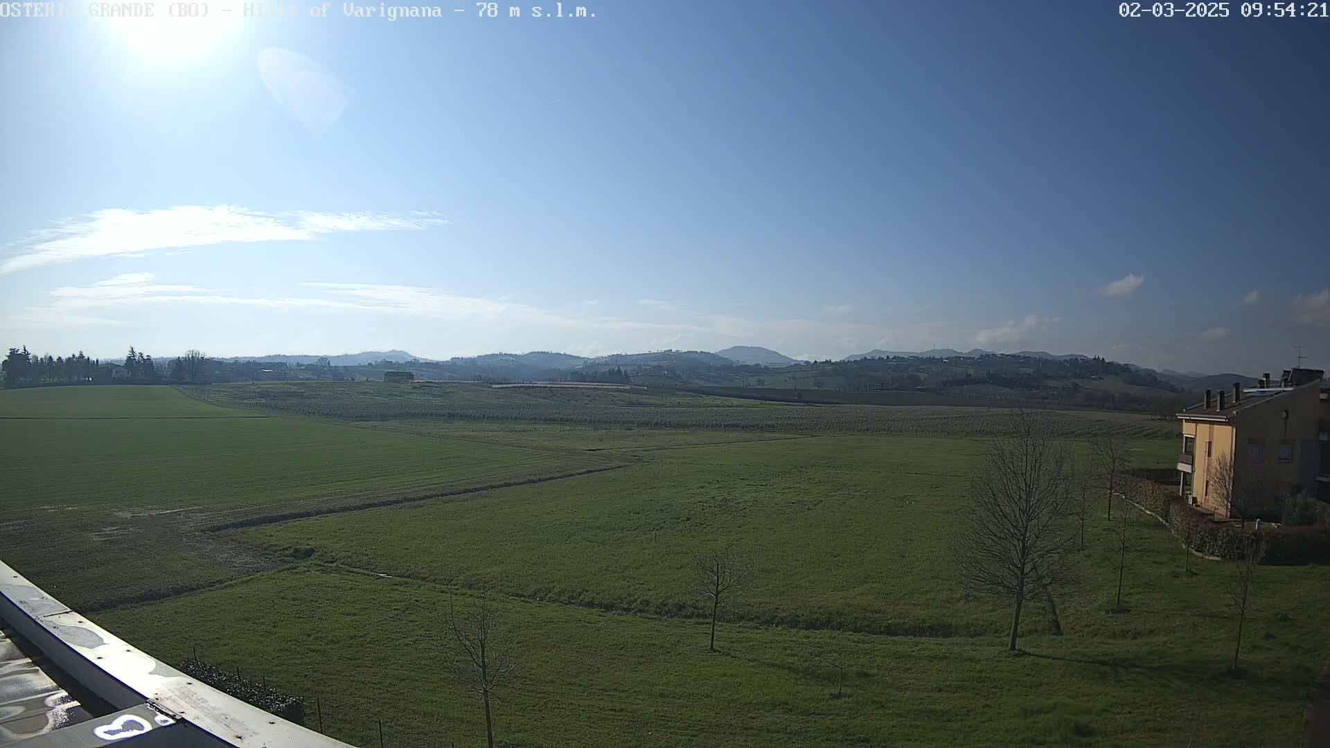 A mostly sunny day reveals a vast expanse of green fields, dotted with a few trees and a distant building, all set against a backdrop of rolling hills under a clear blue sky.
