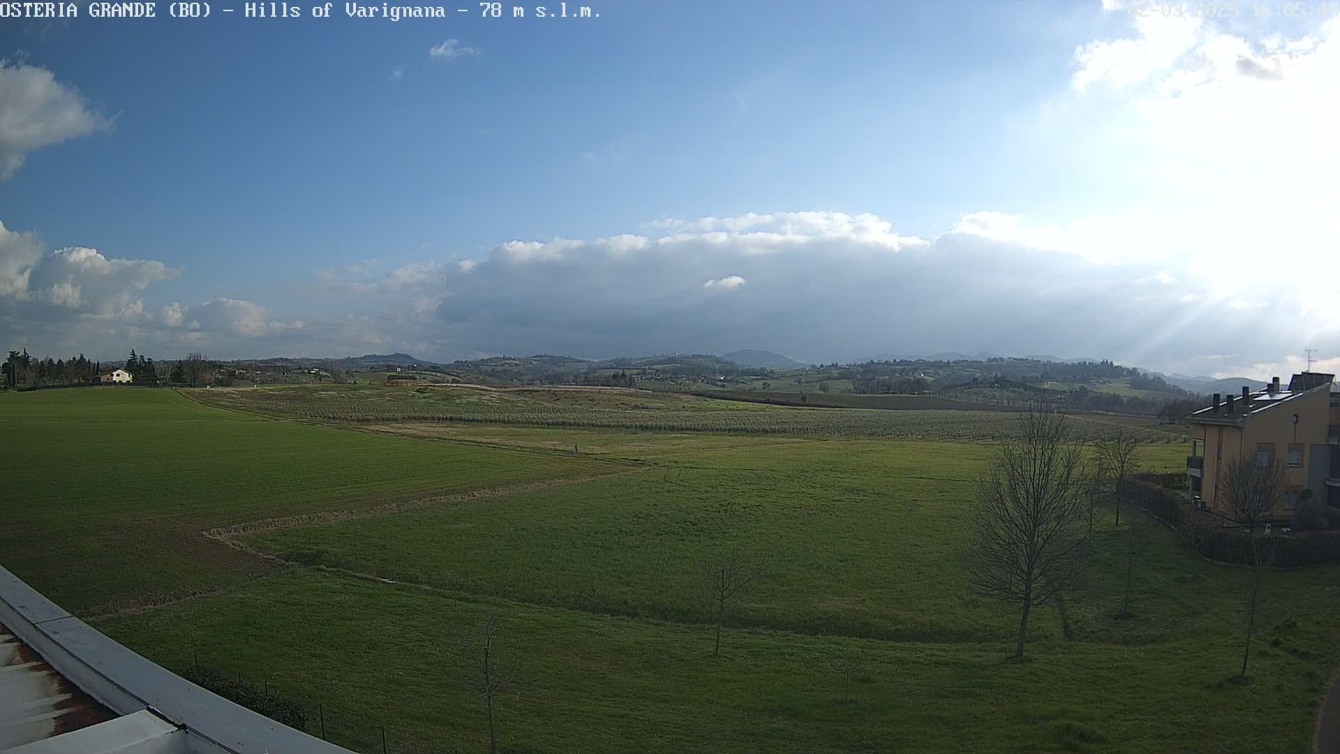 A mostly sunny day with some clouds over a sprawling green field, distant hills, and a few buildings.