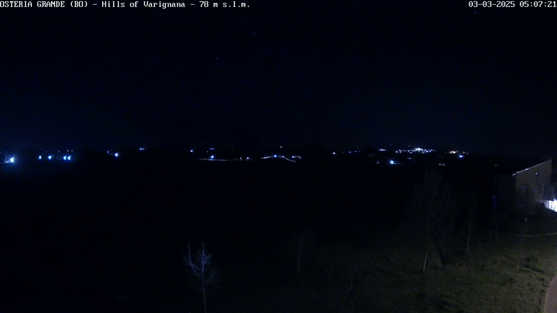 A nighttime view of dimly lit hills and a few buildings in the distance, under a clear, dark sky.