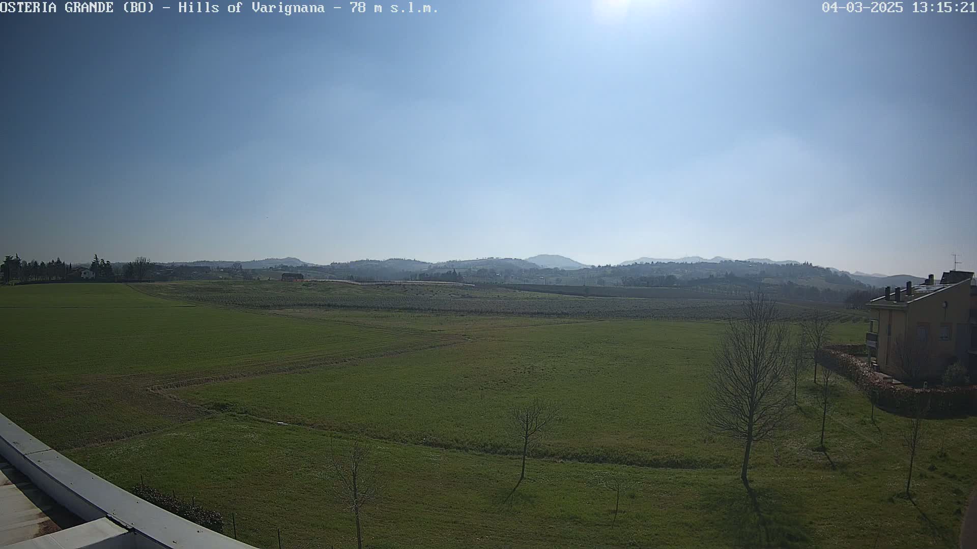 A sunny day reveals a vast, green field stretching towards distant, hazy hills, with a few leafless trees and a building visible in the foreground.