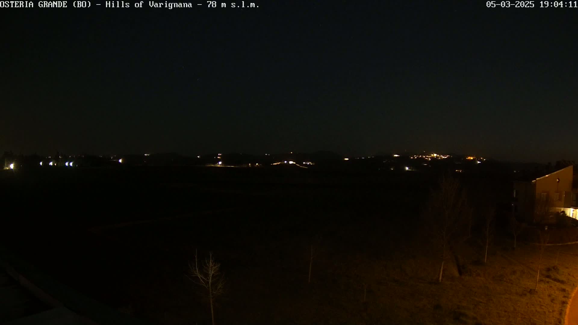 A dark, nighttime view of sparsely lit hills and a few buildings, under a clear sky.