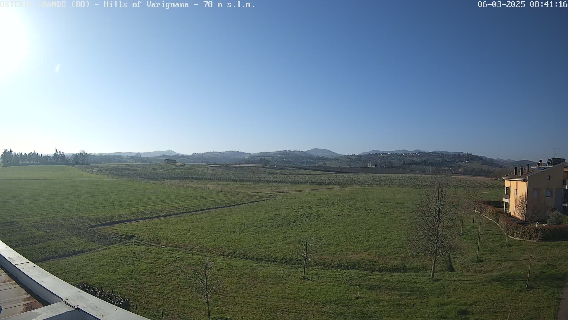 A wide, expansive view of a green field under a clear blue sky, with distant hills and a single yellow building visible in the bottom right corner.