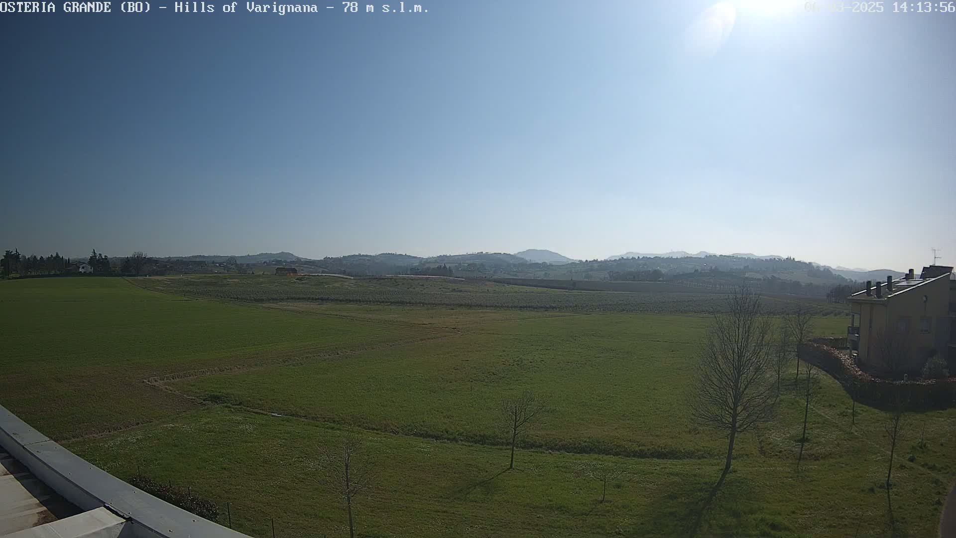 A sunny day reveals a vast expanse of green fields, dotted with a few bare trees and a distant building, all set against a backdrop of rolling hills under a clear blue sky.