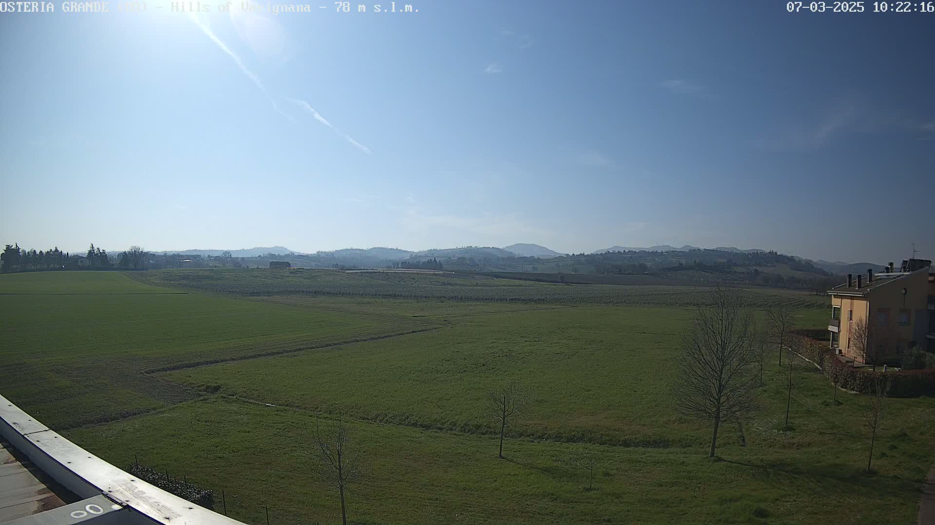 A wide, flat expanse of green fields stretches to rolling hills under a clear, bright blue sky.