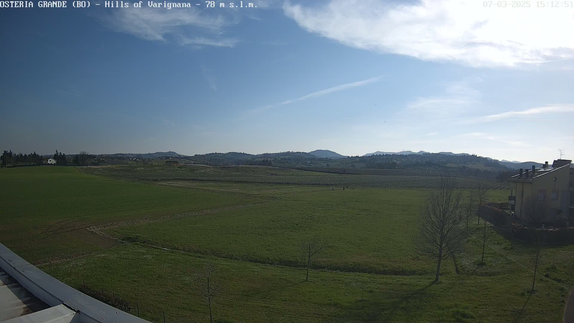 A sprawling green field stretches towards distant hills under a mostly clear, bright sky.