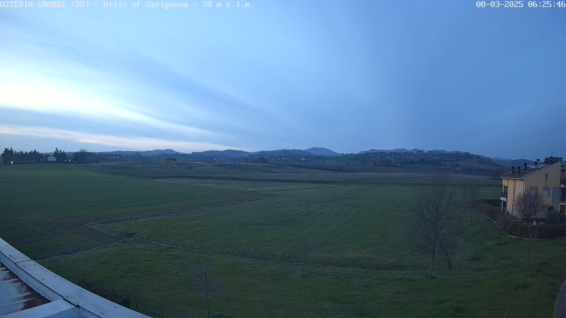 A wide, flat expanse of green farmland stretches towards distant, softly lit hills under a twilight sky.