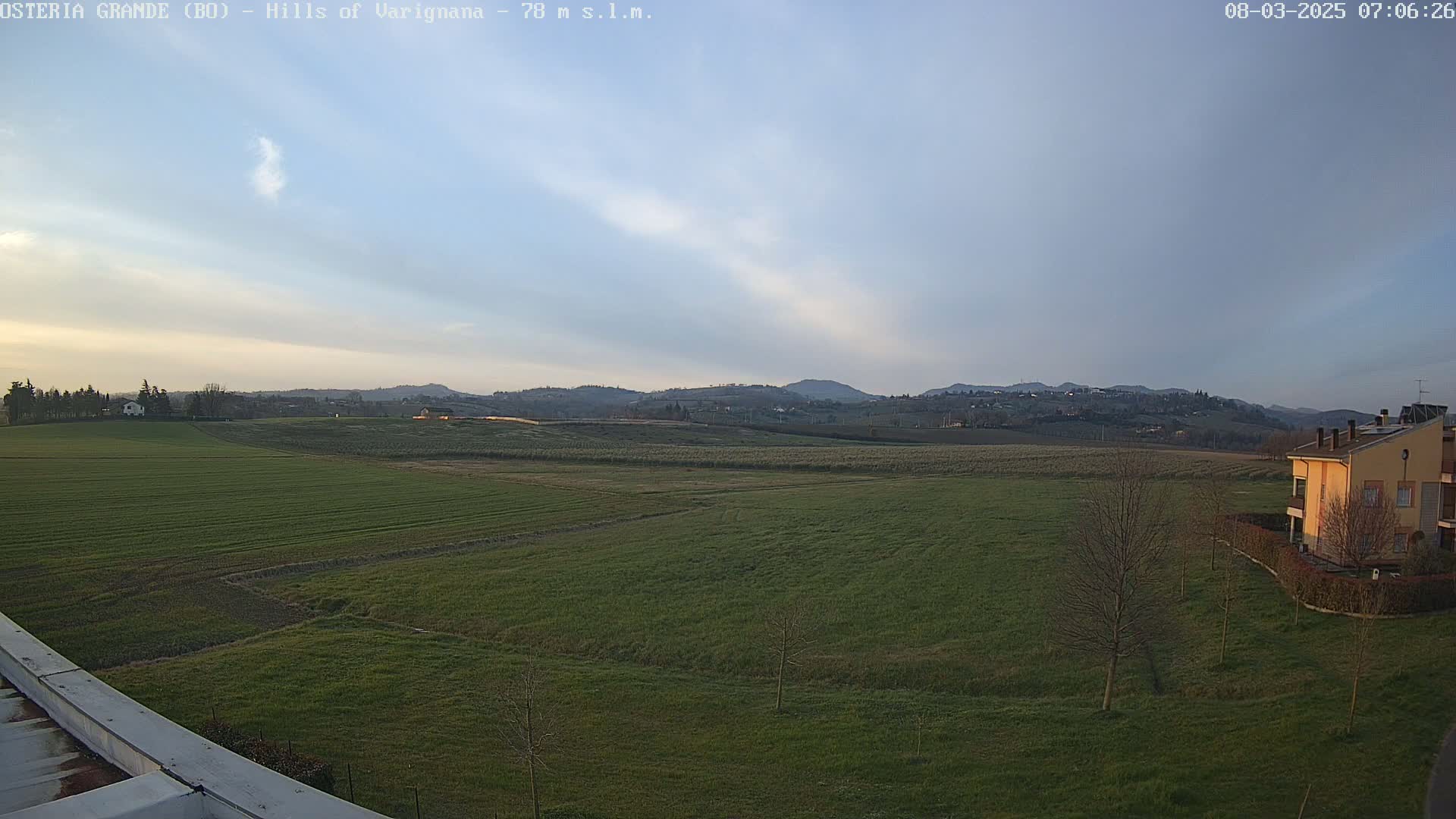 A sprawling green field stretches out under a pale blue sky with thin, wispy clouds,  backed by distant rolling hills and a few buildings.