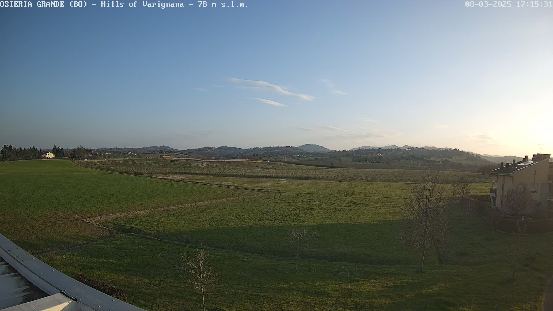A clear, sunny day reveals a sprawling green field, dotted with a few buildings, leading to rolling hills in the distance.