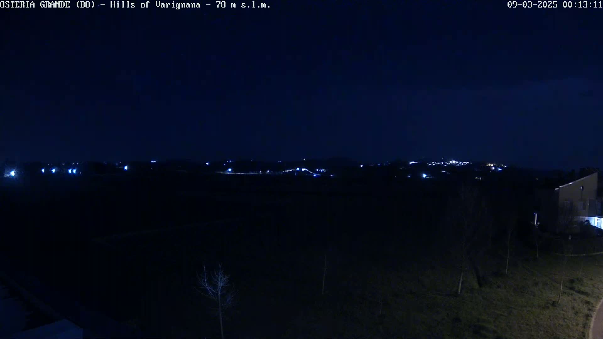 A nighttime long shot shows a dark, mostly treeless landscape with a distant town’s lights visible on a hill under a clear, dark sky.