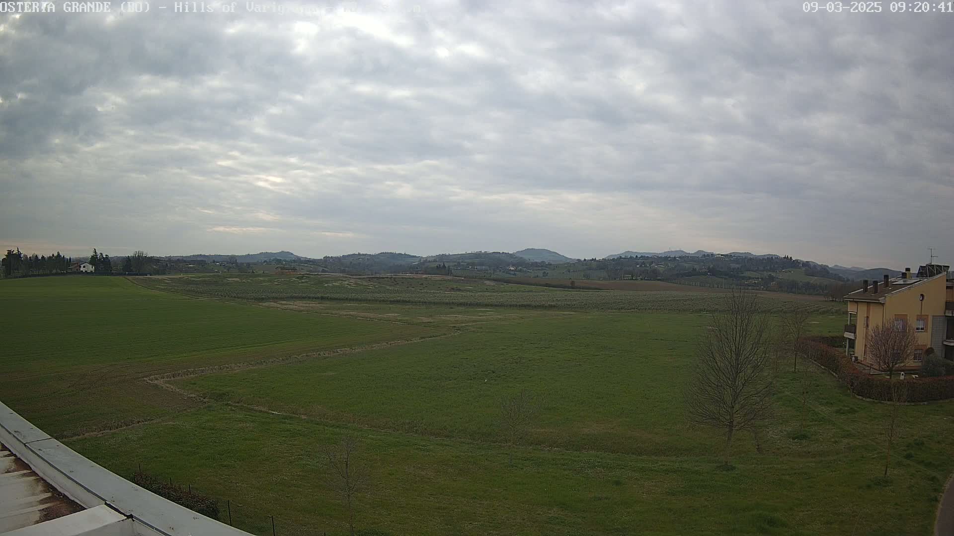 A wide, green field stretches towards a line of low hills under a mostly cloudy sky.