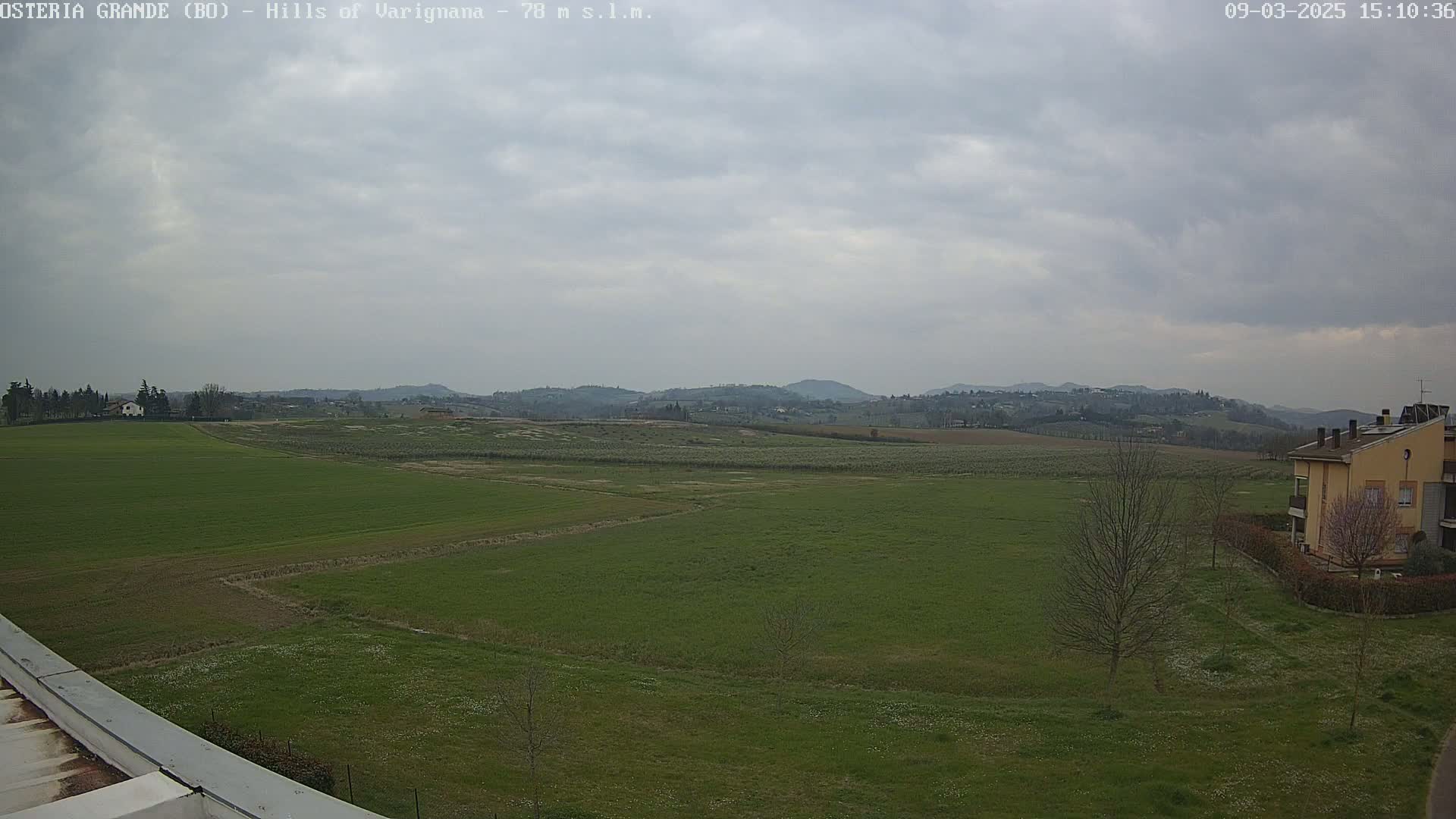 A wide, flat expanse of green fields stretches towards distant, rolling hills under a cloudy sky.