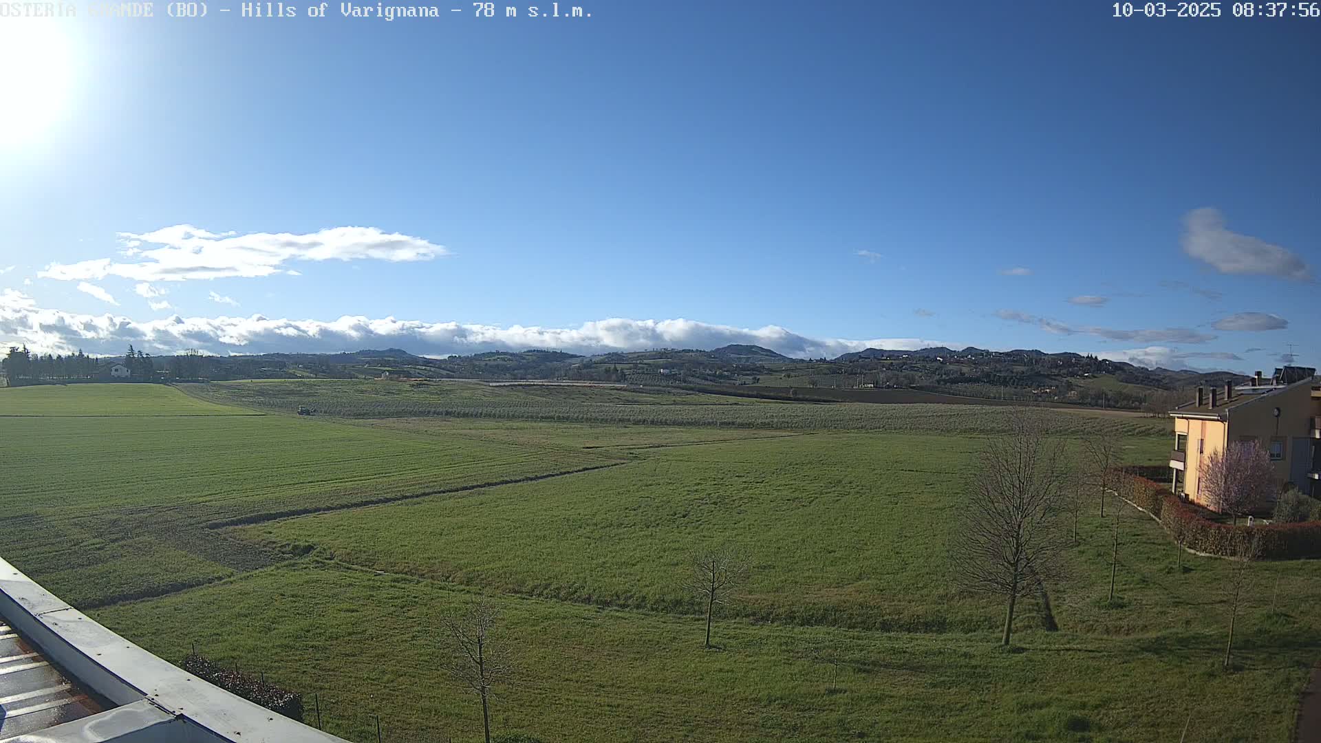 A wide, flat expanse of green farmland stretches to rolling hills under a mostly clear blue sky with scattered clouds.