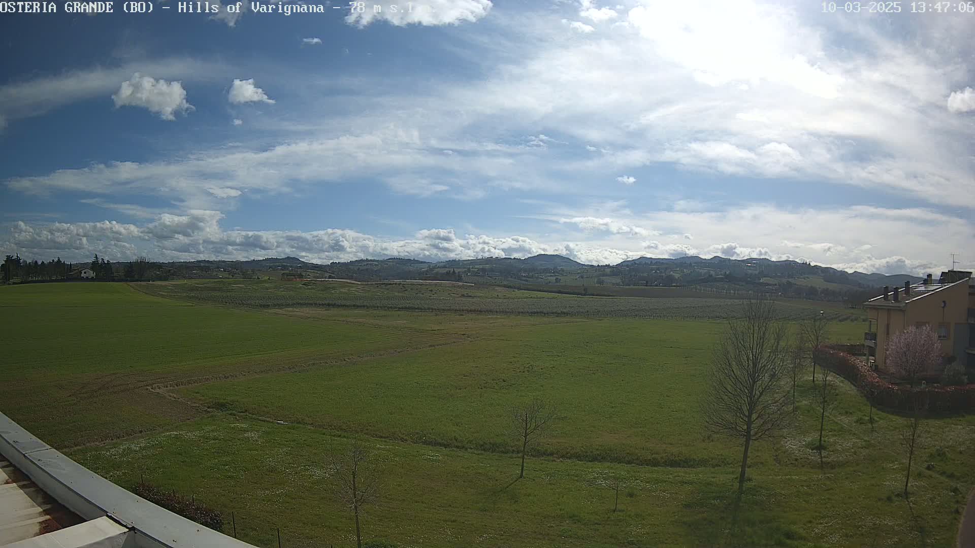 A partly cloudy sky overlooks a vast expanse of green fields, dotted with a few trees and a building in the distance, under a bright sun.