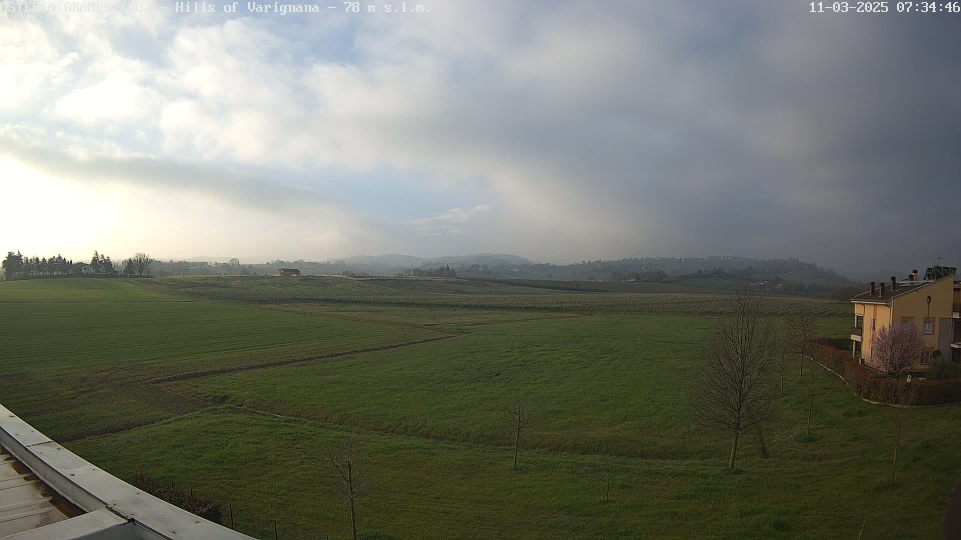 A sprawling green field stretches towards rolling hills under a mostly cloudy sky.