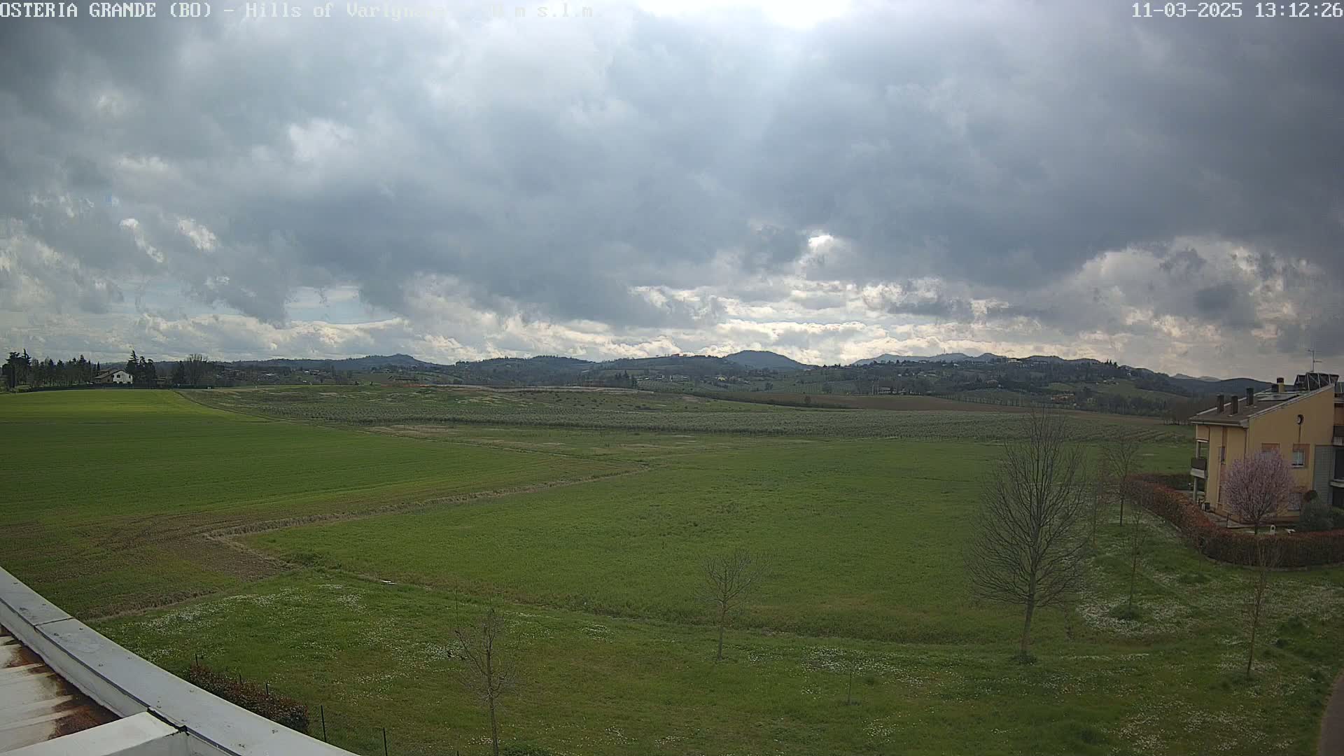 A mostly cloudy sky over a wide expanse of green fields, dotted with a few buildings and trees, and distant hills in the background.