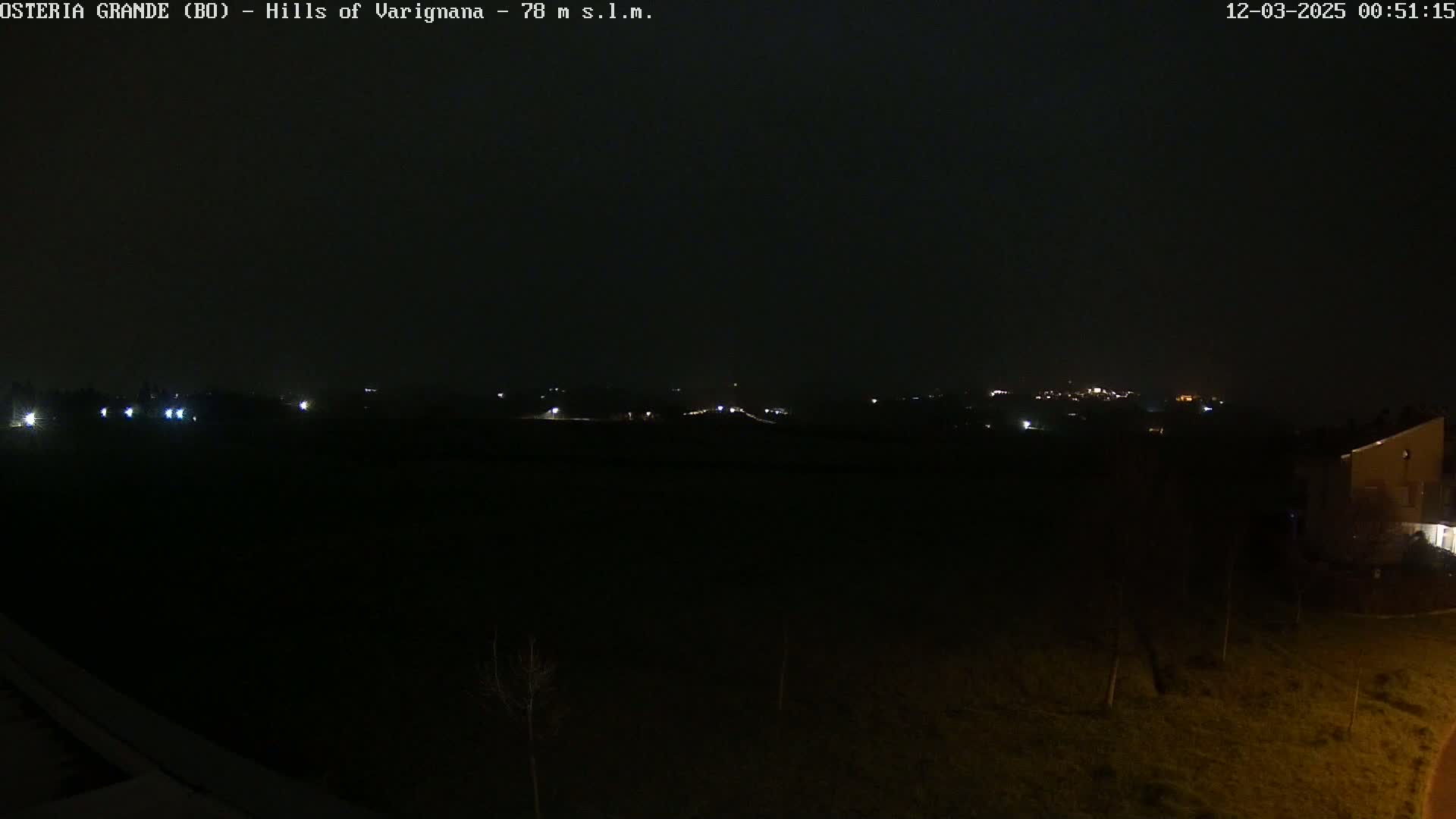 A dark, nighttime scene shows a flat, grassy area with a few distant lights on a hill in the background, under a dark, possibly overcast sky.
