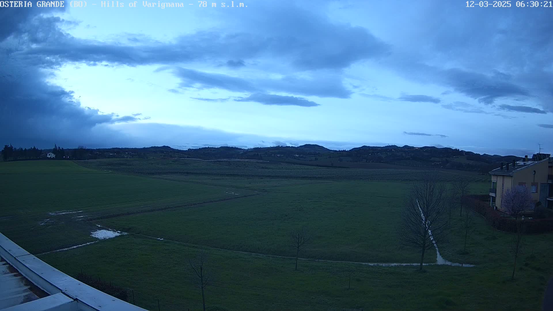 A mostly cloudy, pre-dawn sky over a flat, green field with a few buildings and trees in the distance.