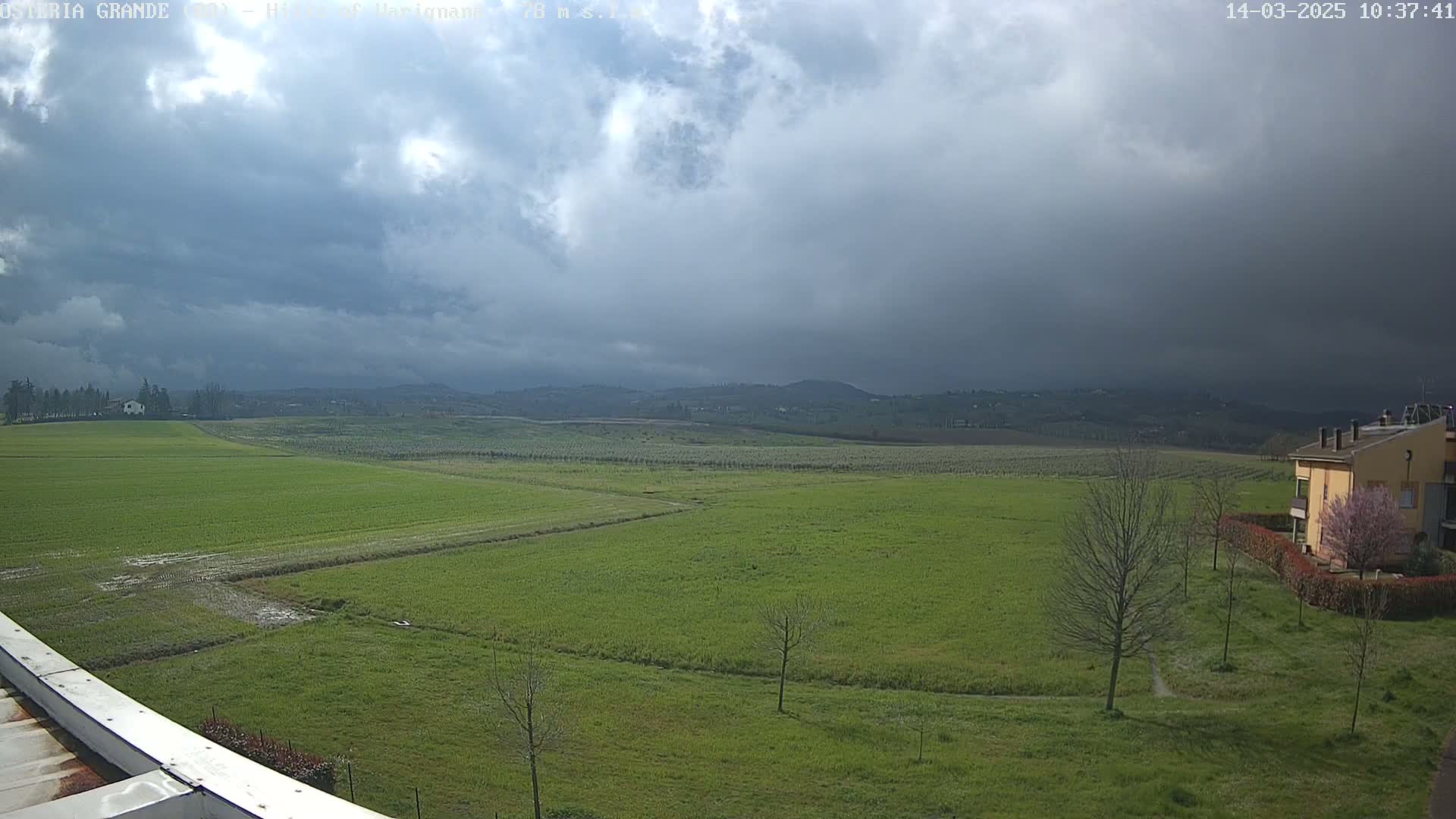 A sprawling green field stretches under a cloudy sky, with a few buildings and trees visible in the distance.