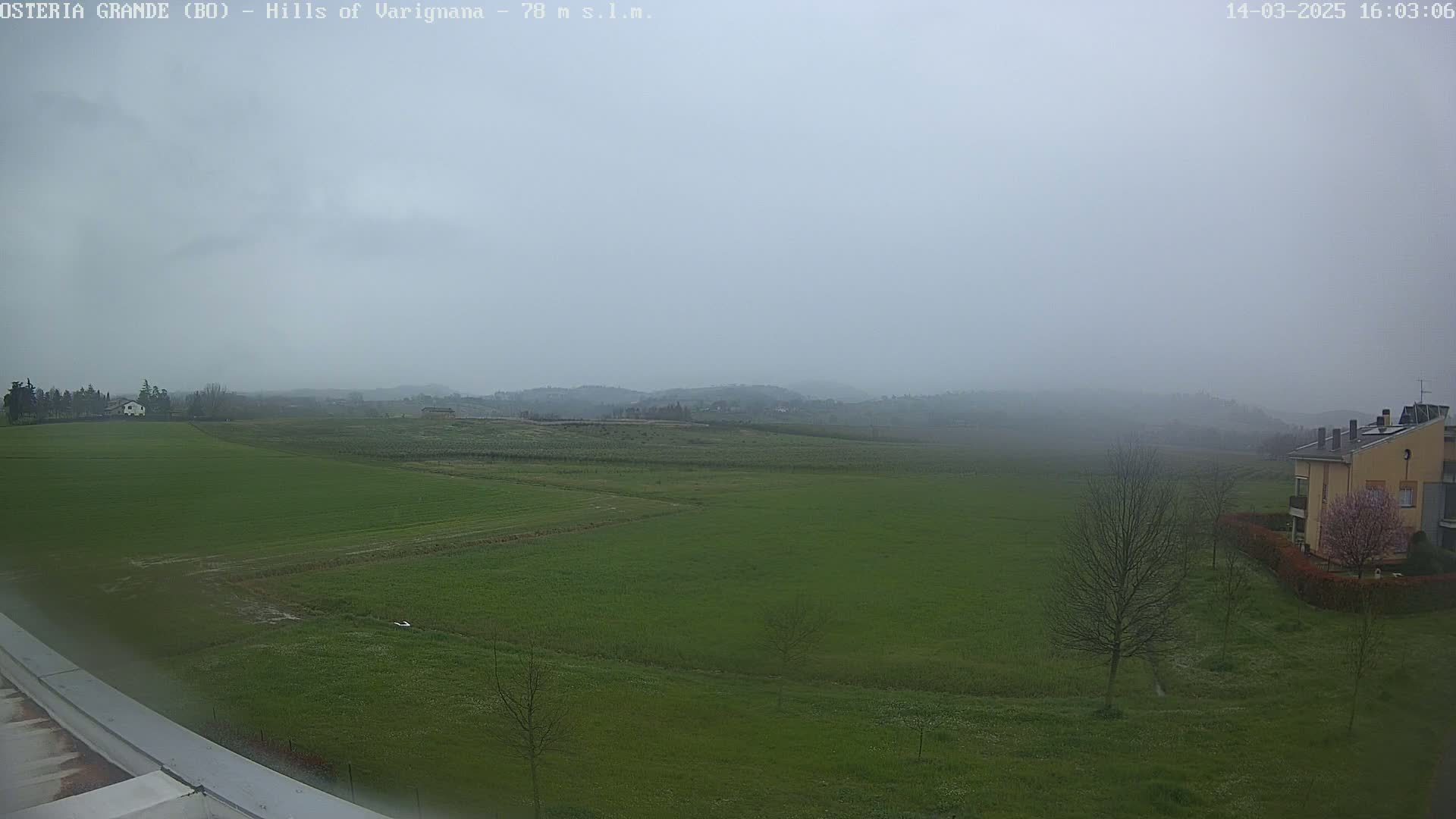 A foggy, overcast view of a flat, green field, a few buildings, and distant hills.