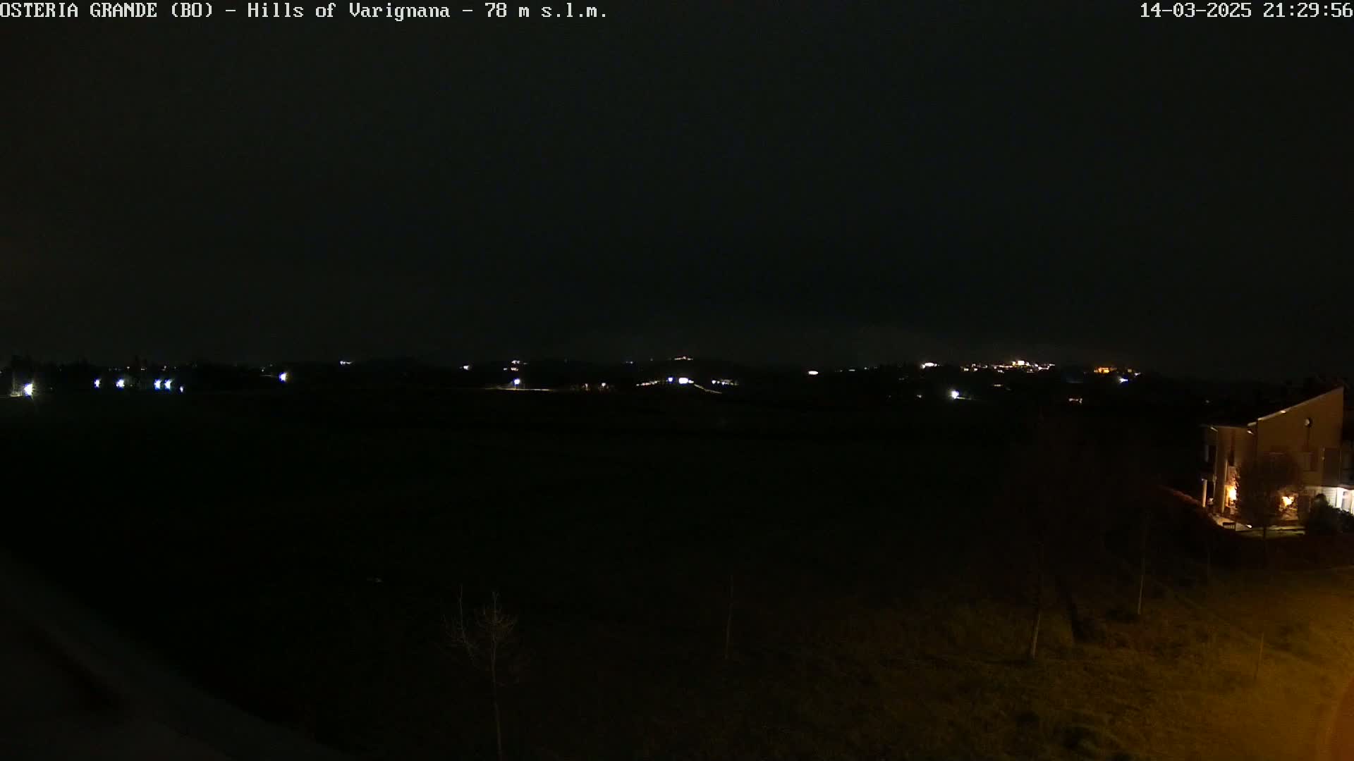 A dark, nighttime view of a rural landscape with a few distant lights and a building in the lower right corner.