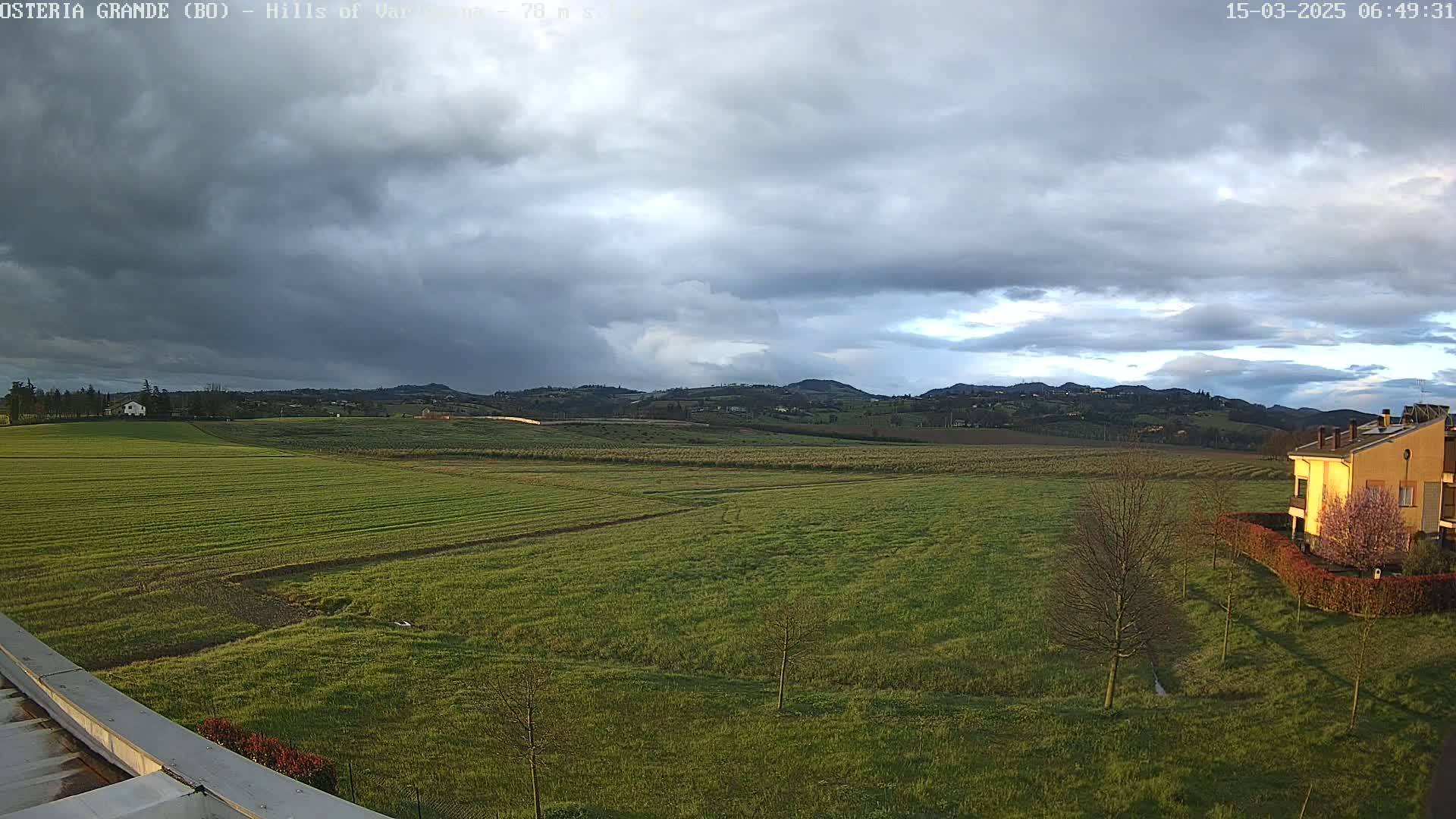 A vast, flat green field stretches toward rolling hills under a mostly cloudy sky.