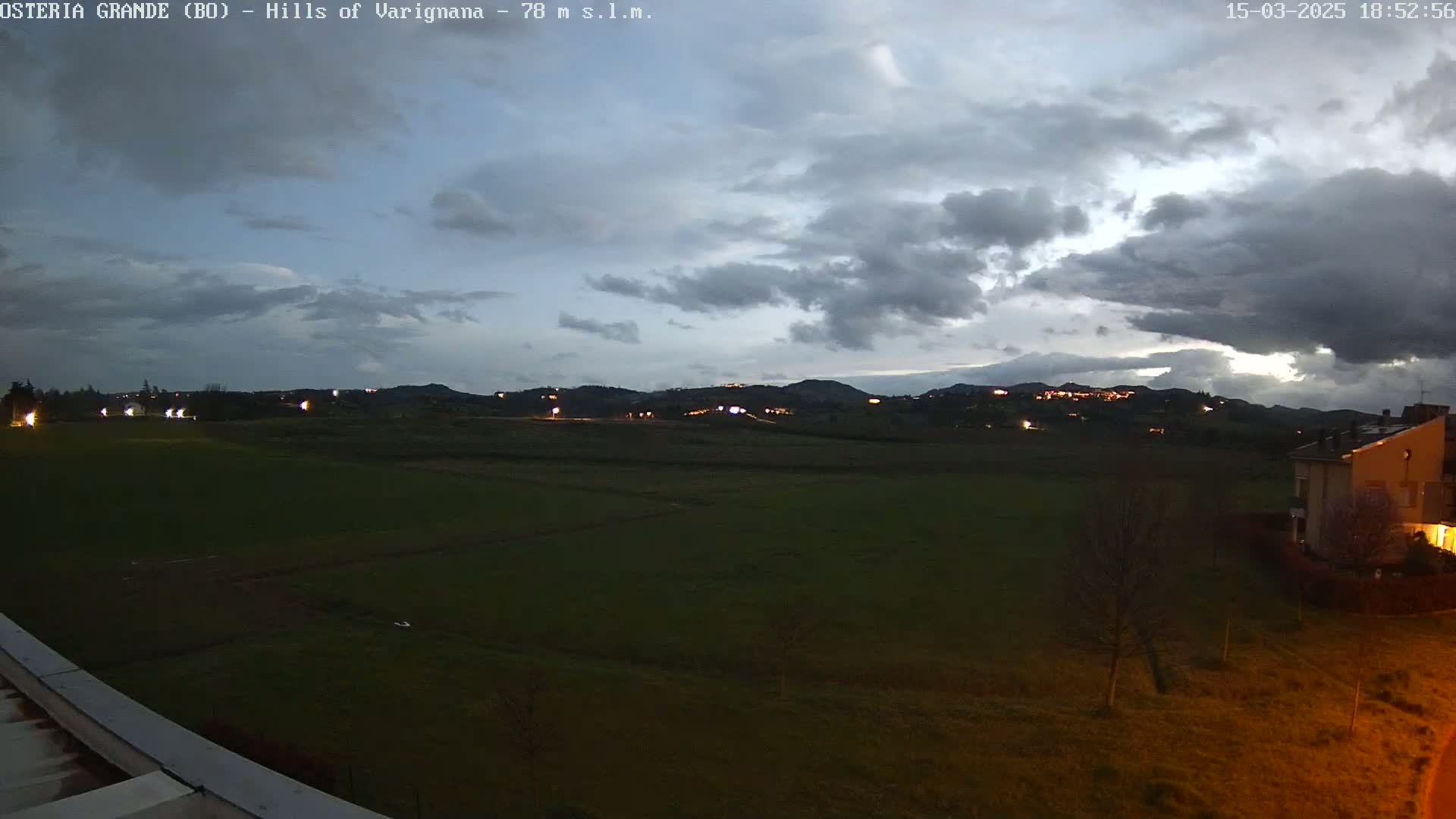 A view of a dark green field at dusk under a cloudy sky, with distant hills illuminated by lights and a building visible in the lower right corner.