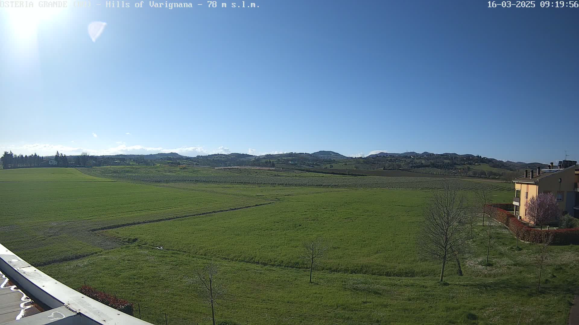 A sunny day reveals a sprawling green field, bordered by trees and a building, with rolling hills in the background under a clear blue sky.