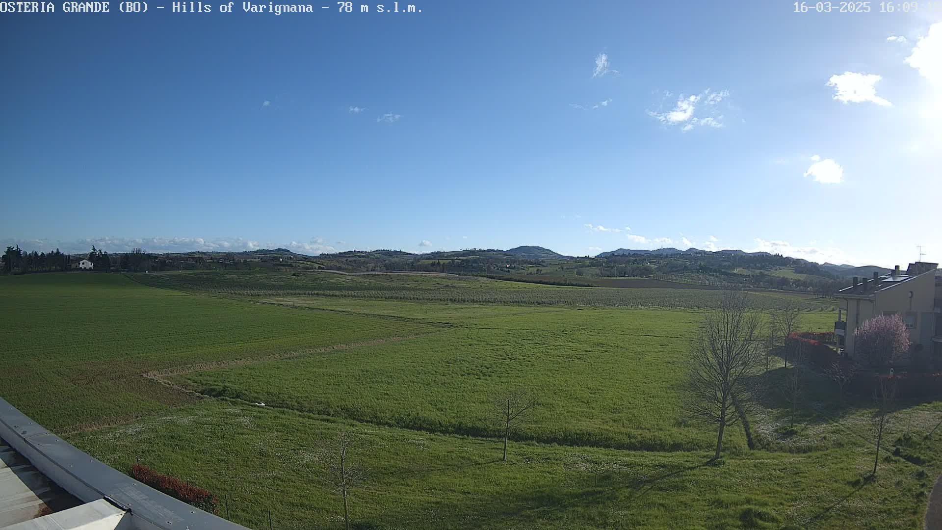 A sprawling green field stretches towards rolling hills under a mostly clear blue sky with a few scattered clouds.