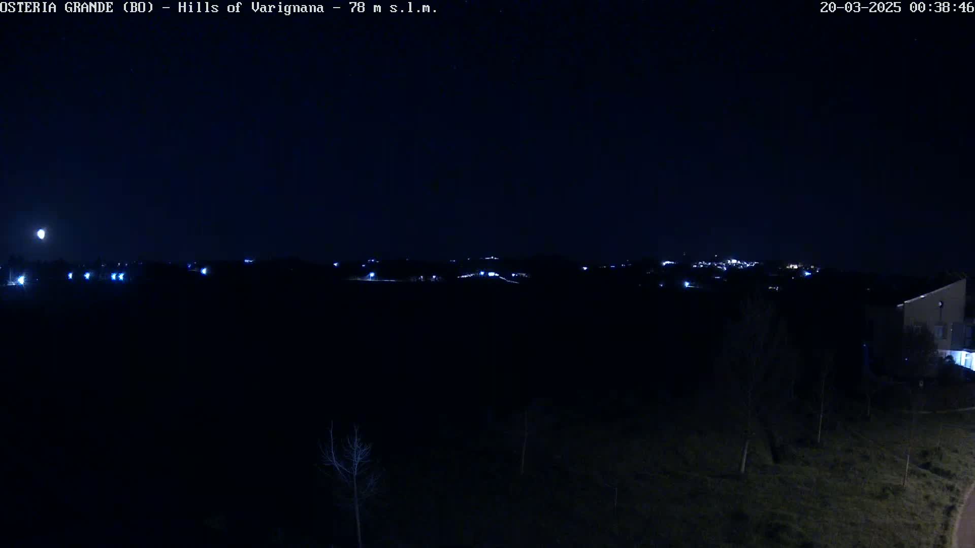 A nighttime view of a dark landscape with a few distant lights visible in the hills under a mostly clear, moonlit sky.