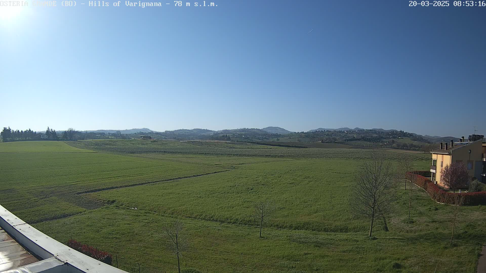 A vast, green field stretches towards rolling hills under a clear blue sky.