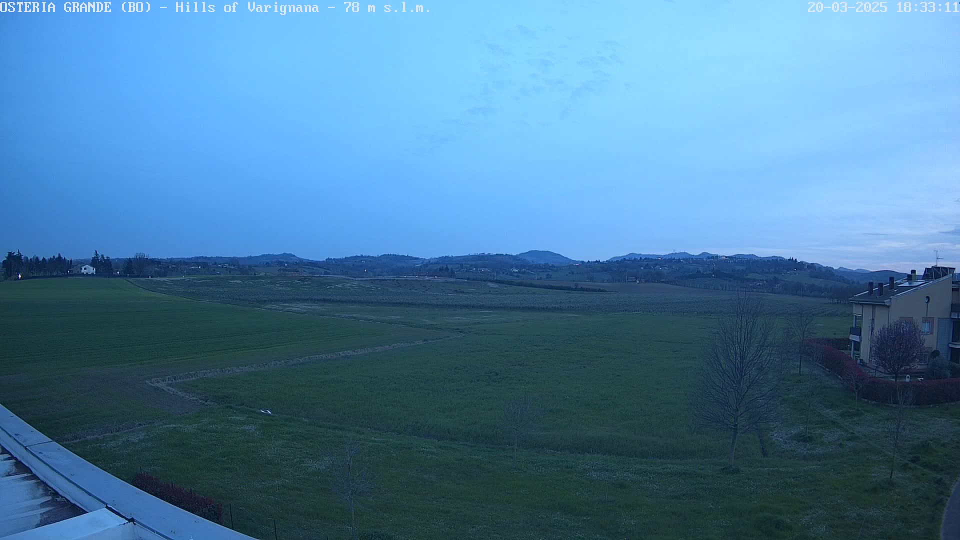 A twilight view of rolling green fields, a few buildings, and distant hills under a clear, pale blue sky.