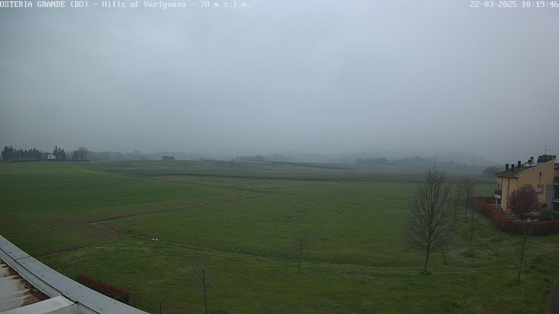 A hazy, overcast view shows a sprawling expanse of green fields, dotted with a few trees and buildings in the distance, and a building closer to the foreground.