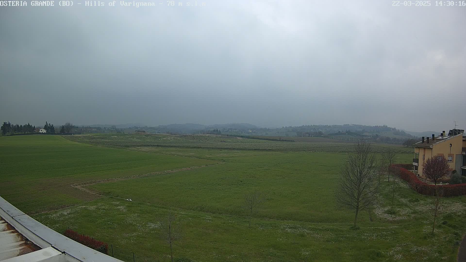 A sprawling green field stretches out under an overcast sky, with a few buildings visible in the distance and closer to the viewer.