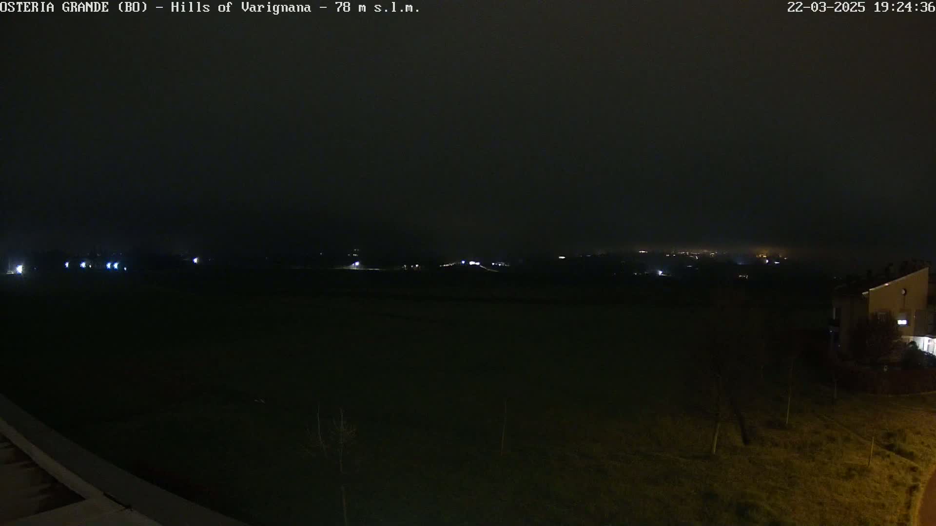 A nighttime view of a dark, flat field with a few distant lights and a building visible on the right, under a foggy sky.