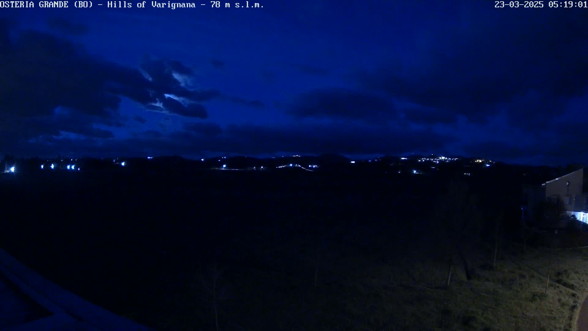 A nighttime view of dimly lit hills and a few buildings under a dark blue, cloudy sky.