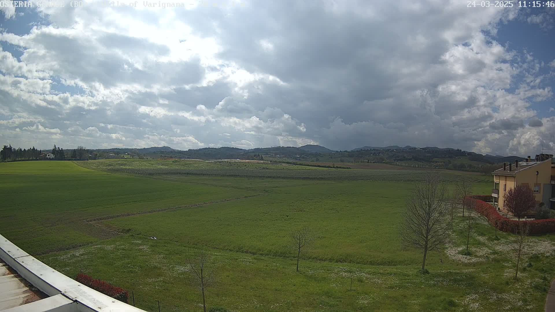 A partly cloudy sky overlooks a vast expanse of green farmland, dotted with a few trees and a building in the distance.