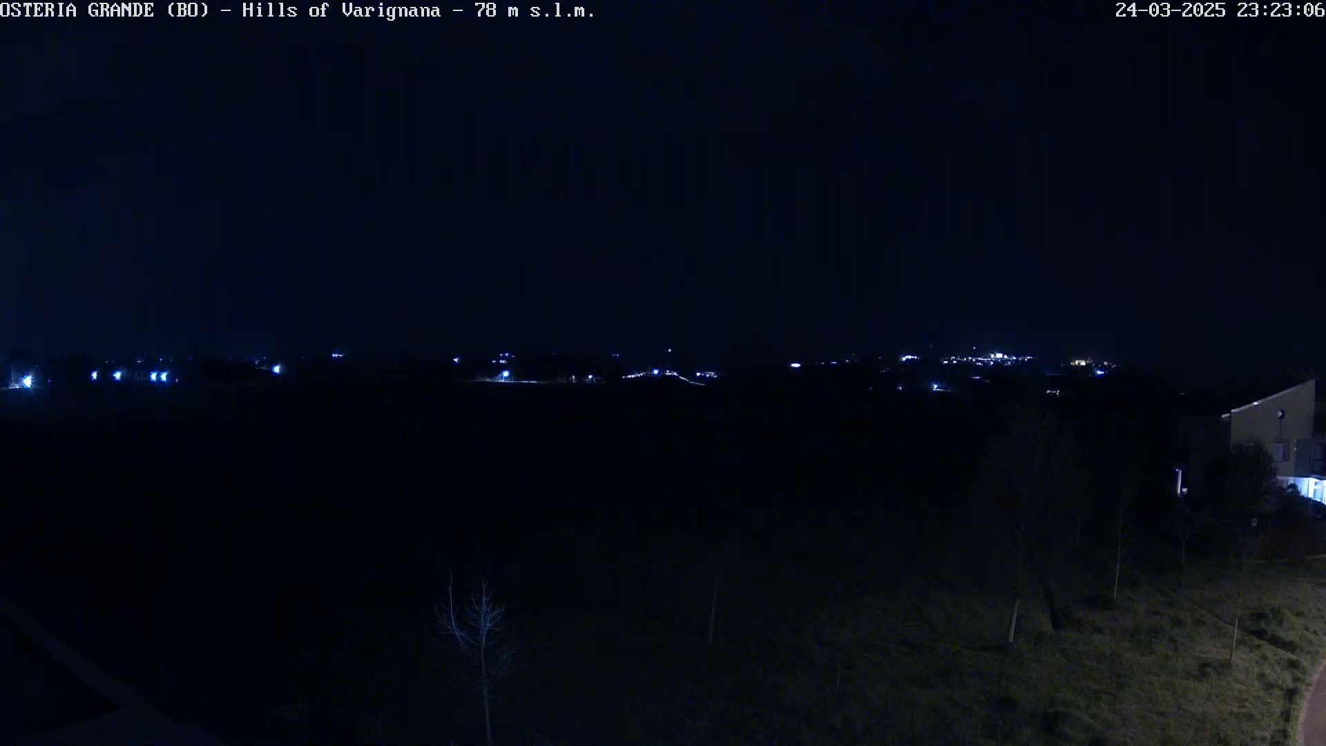 A nighttime view of a dark landscape with a distant line of lights and a building partially visible in the lower right corner.